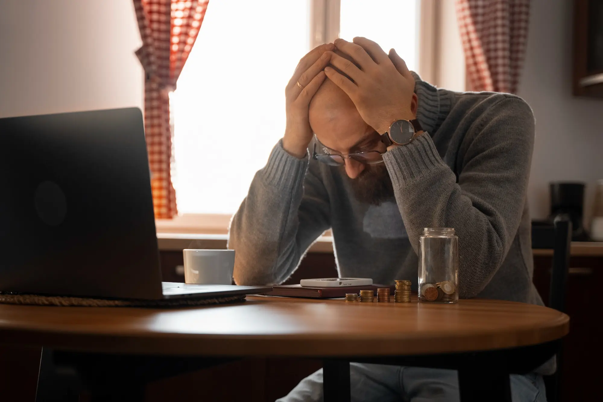 Stressed man holding his head at a desk with a laptop and glass of water.